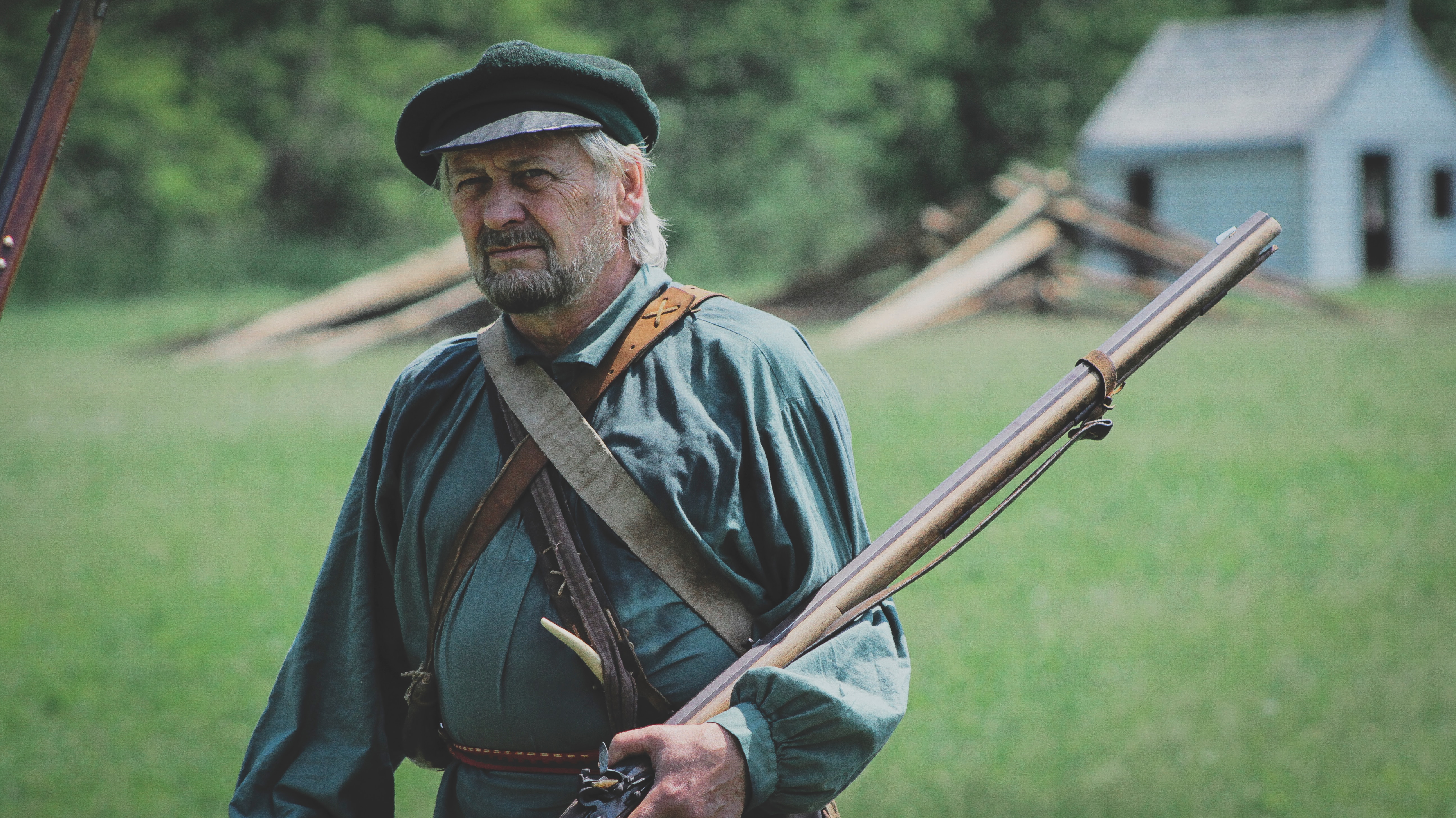 man in colonial farmers' garb, holding  long rifle in the crook of his arm; in the background, a split rail fence and small pale blue shed